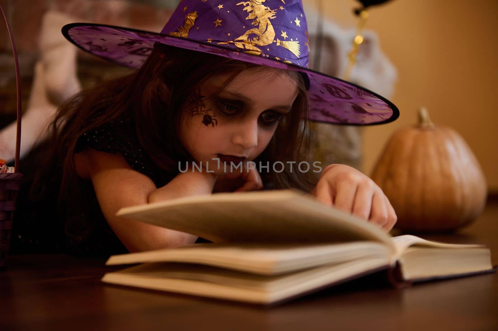 Close-up little girl with a face art make-up for Halloween, dressed as a witch, enchantress in wizard hat reading sorcery and spell magic book surrounded by pumpkins and festive gothic atmosphere