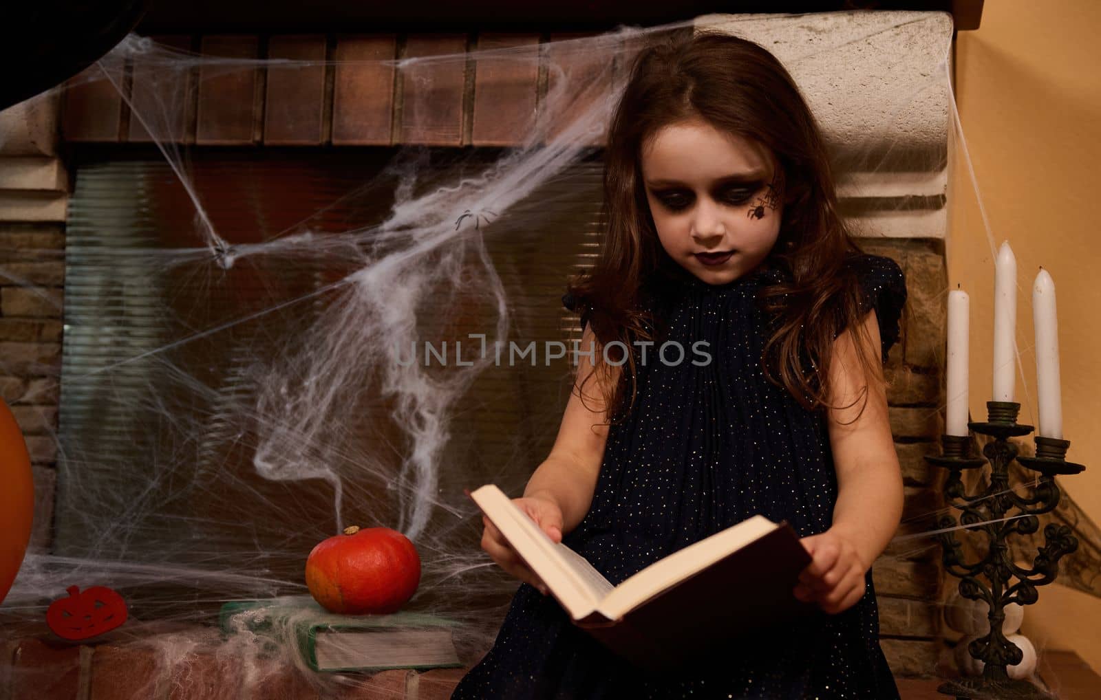 Adorable gothic stylish child girl with a book of spells, sitting by a cobweb-covered fireplace and candles on a candlestick holder, learning enchentery, sorcery while celebration the Halloween night