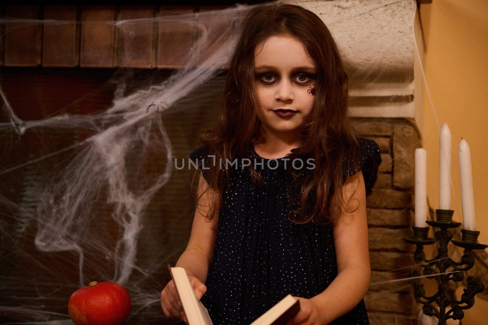 Caucasian gothic child with a spider paint on her cheek, little girl sorceress, witch, enchantress with a spell book, sits against a cobweb-covered fireplace and pumpkins, looking at camera. Halloween