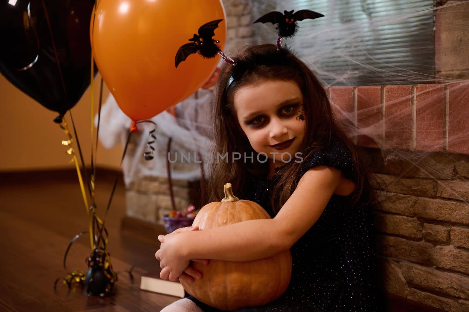 A little sorceress sits by a fireplace with cobwebs and orange and black balloons, hugs Halloween pumpkin Jack-O-Lantern by artgf