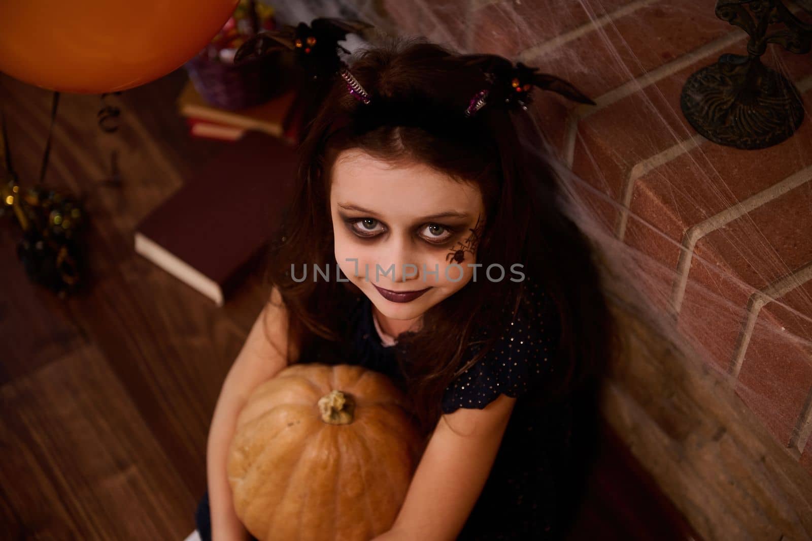 Top view Adorable gothic child, a little girl witch, enchantress, sorceress with face art makeup for Halloween party, hugging a pumpkin, smiles at camera, surrounded by a sorcery book and spider webs