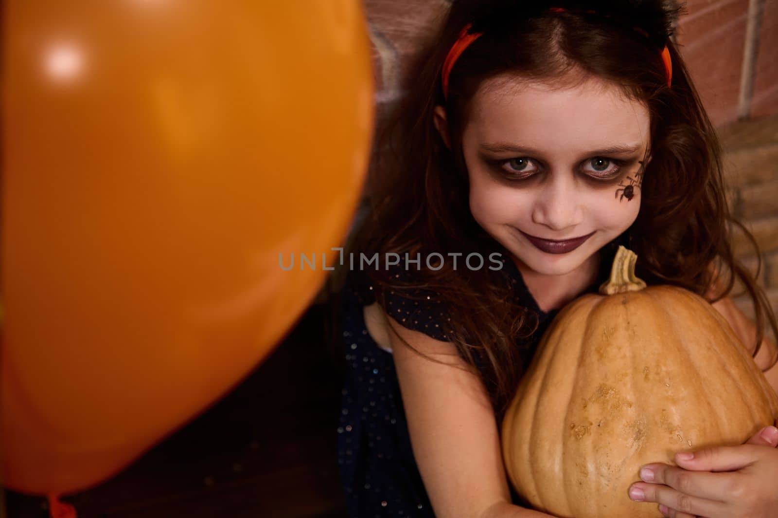 Close-up. Top view of a charming Caucasian little girl with face art makeup - a black spider painted on her cheek, looks like a witch, enchantress hugging pumpkin, smiles at camera. Happy Halloween