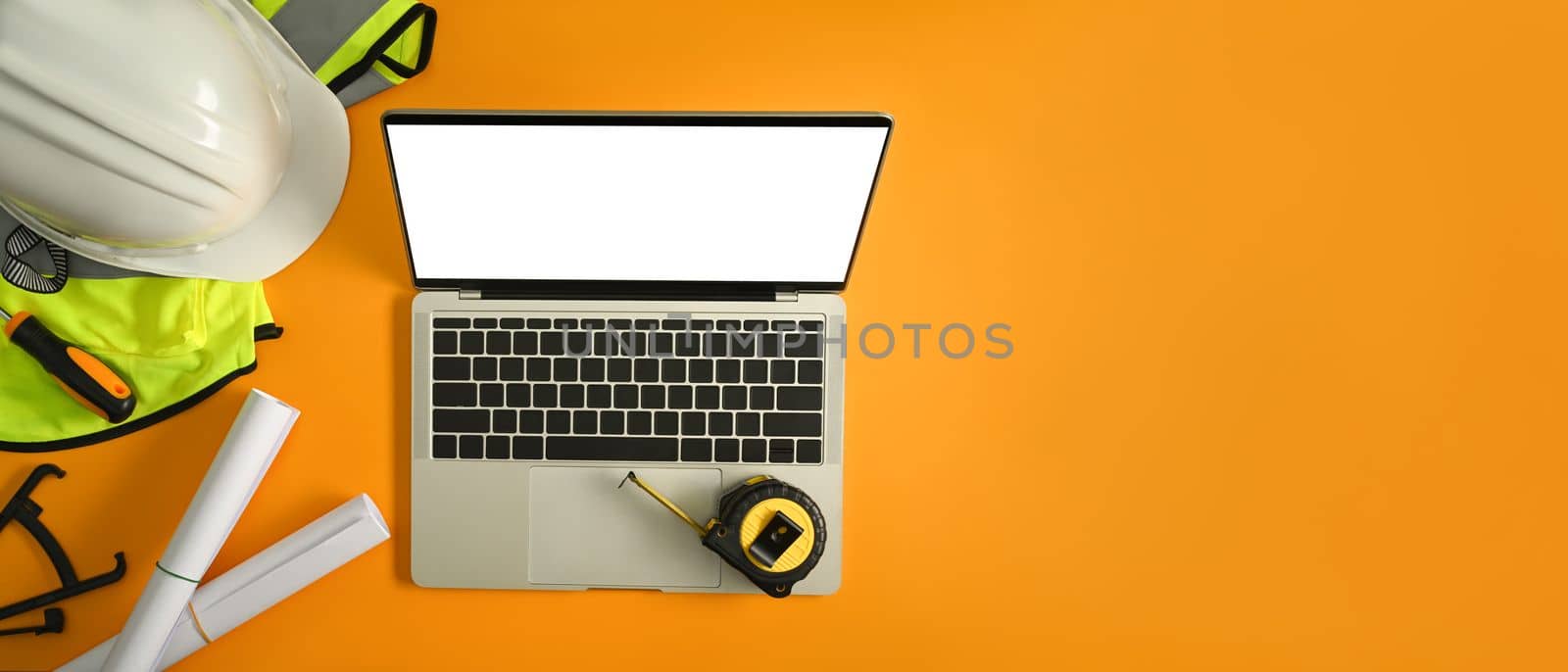 Laptop, yellow vest, safety helmets and blueprints on yellow background. Top view with copy space for your text.