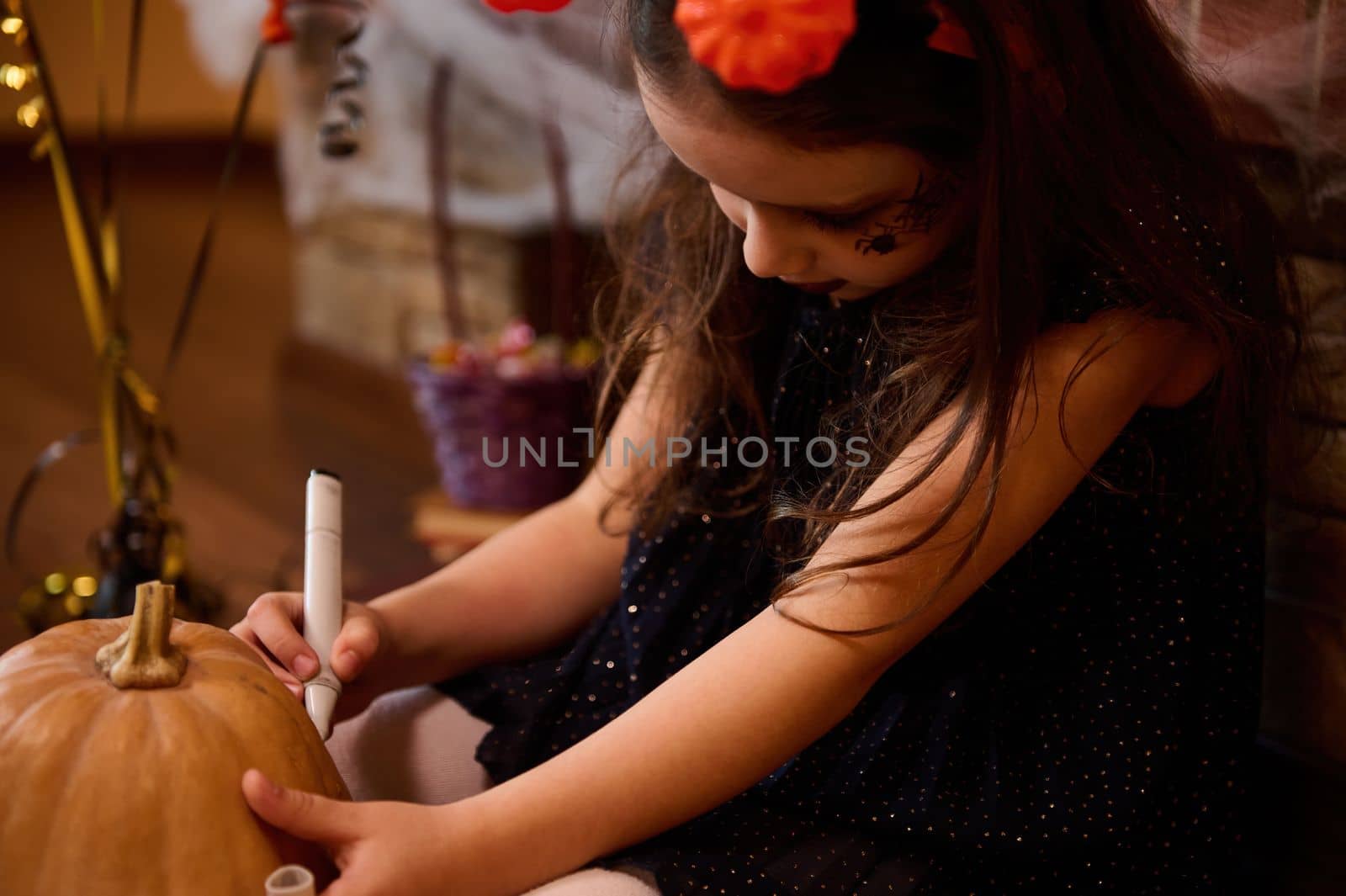 Selective focus on a little girl looking like an enchantress, sitting on the floor surrounded by Halloween treats against a cobweb covered fireplace, holding a pumkin and drawing a Halloween Jack