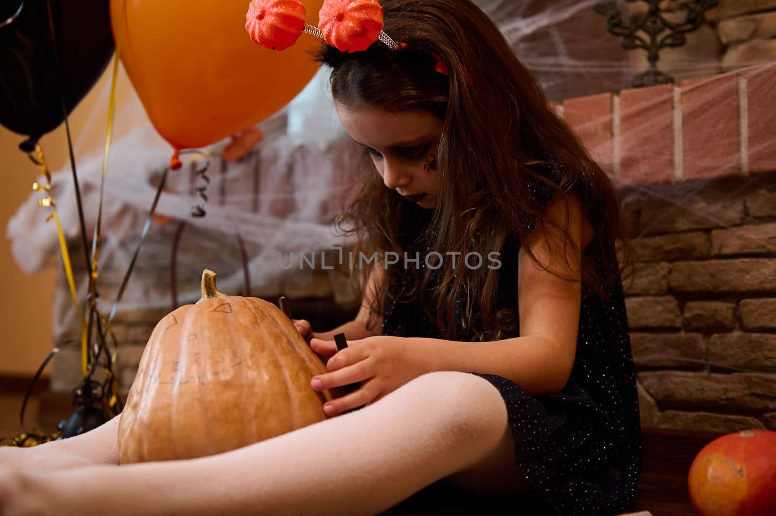 Close-up of an adorable child, a little girl, looking like an enchantress, concentrated on drawing a scary face on a pumpkin, a Jack-O-Lantern, sitting near orange and black air balloons. Halloween