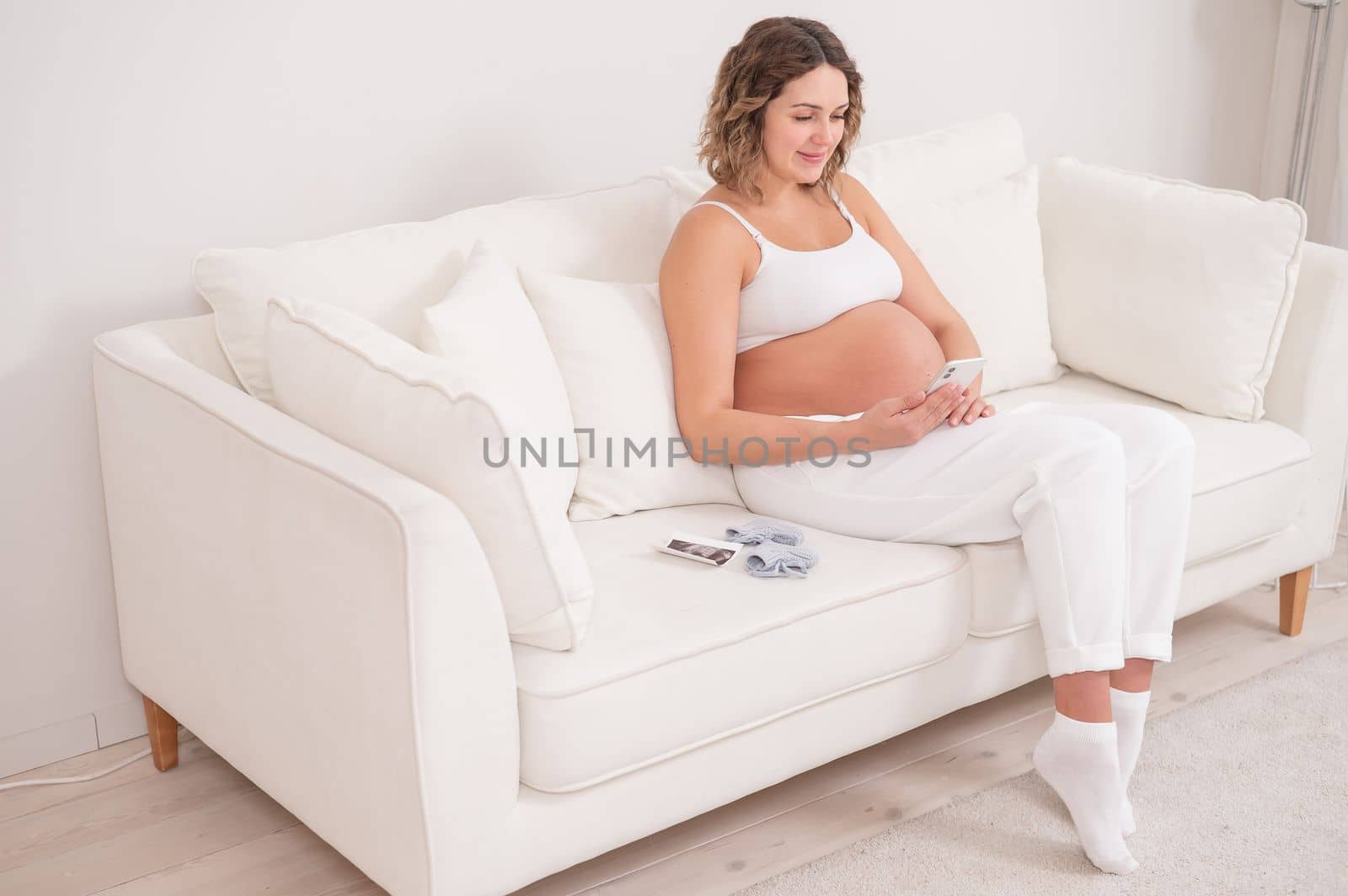 Pregnant woman sits on a white sofa and holds a smartphone. Fetal ultrasonic screening