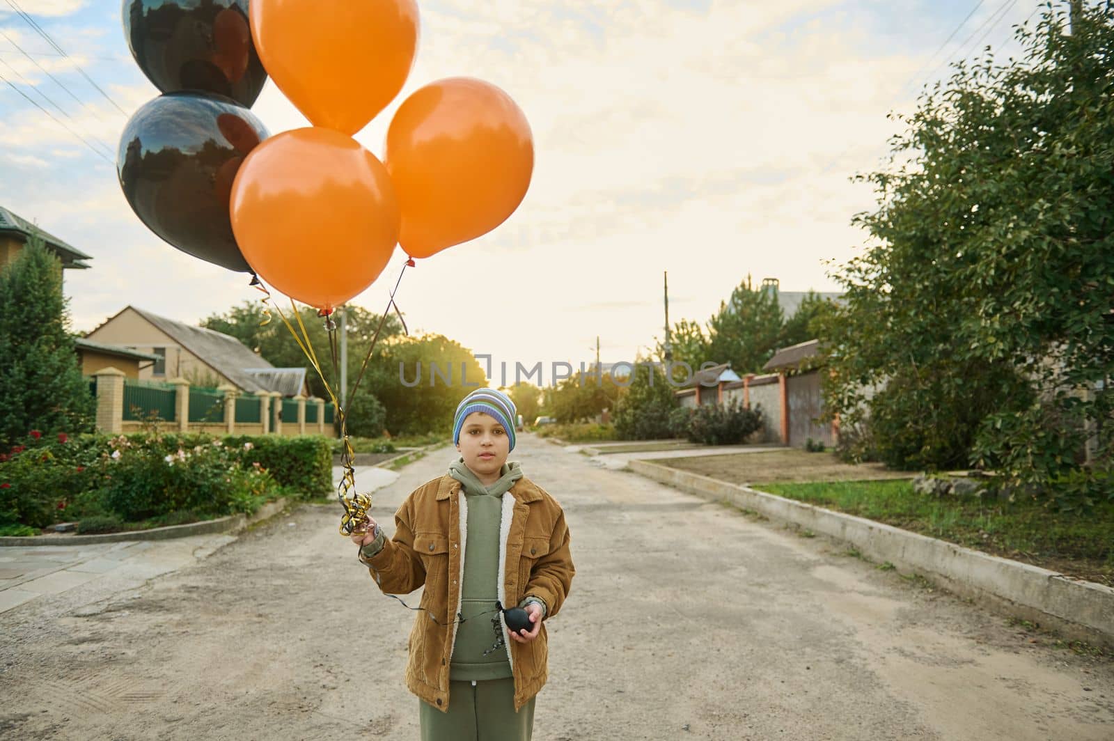 Handsome Caucasian teenage boy in active wear and warm jacket, looking at camera while walking down the village street with air balloons in his hands, at sunset background. Happy Halloween
