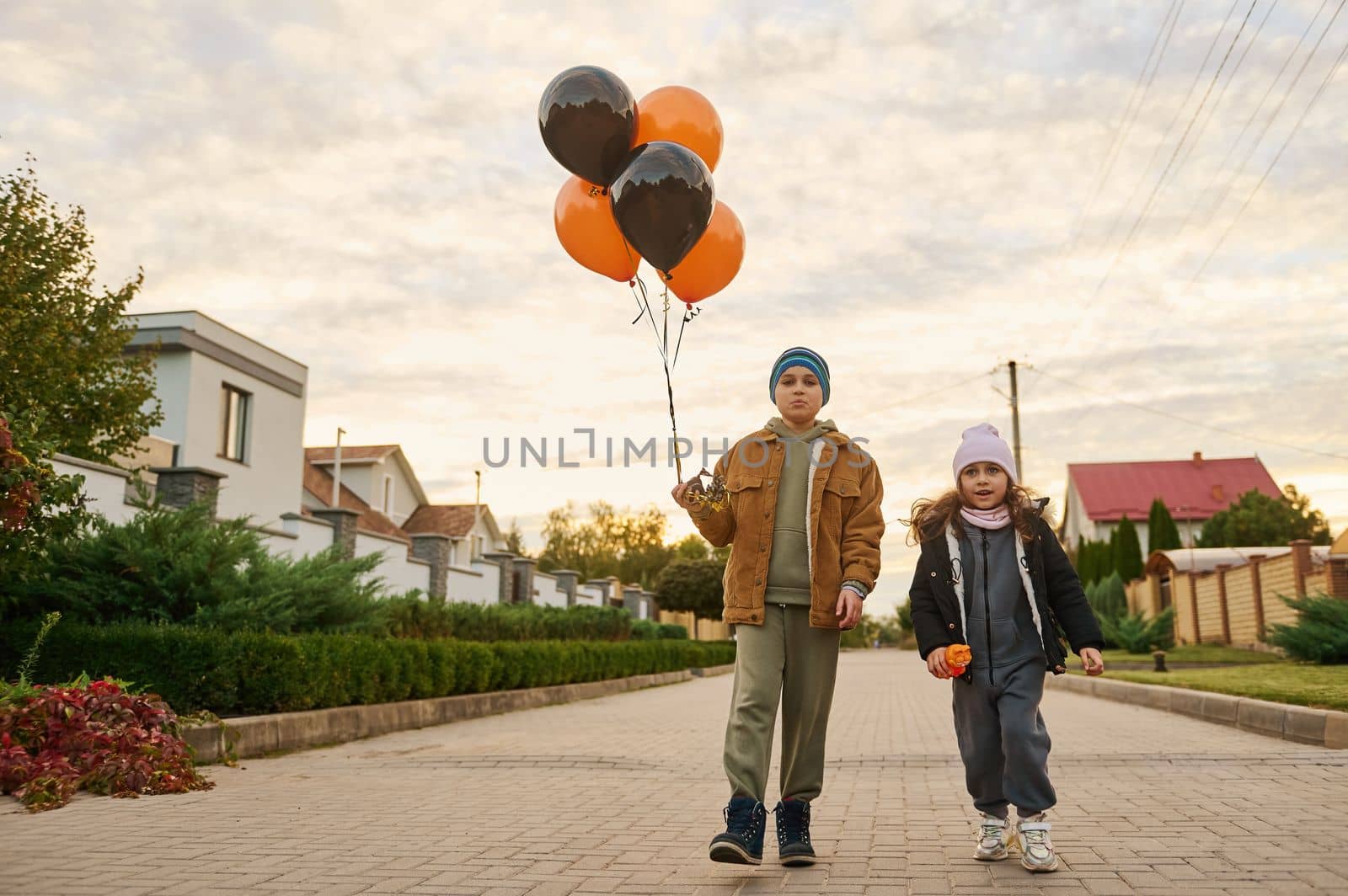 Adorable children, boy and girl, brother and sister with orange black balloons walk down the country street on Halloween by artgf