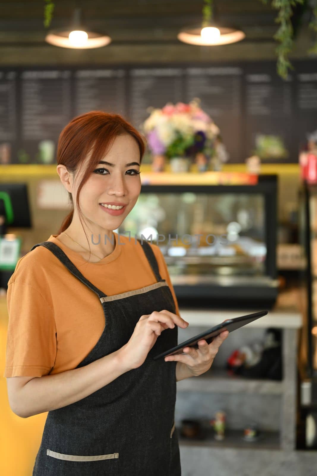 Portrait of smiling female entrepreneur wearing apron standing in front of counter of modern cafe using digital tablet.