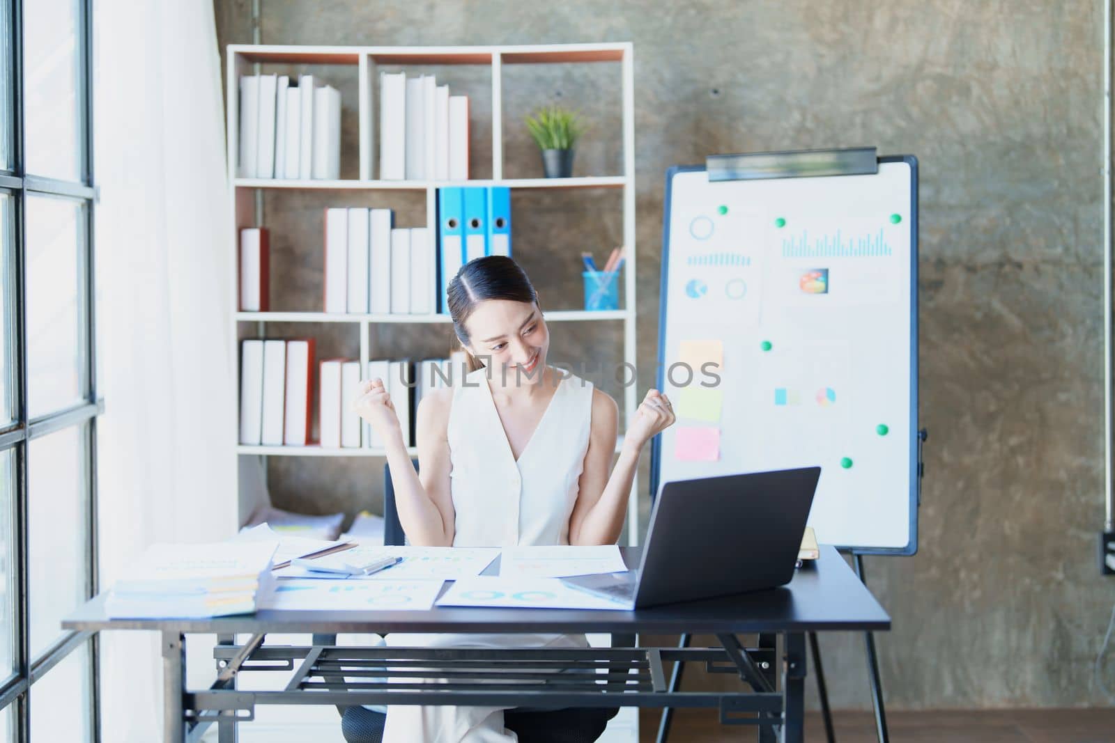 Portrait of a woman business owner showing a happy smiling face as he has successfully invested her business using computers and financial budget documents at work.