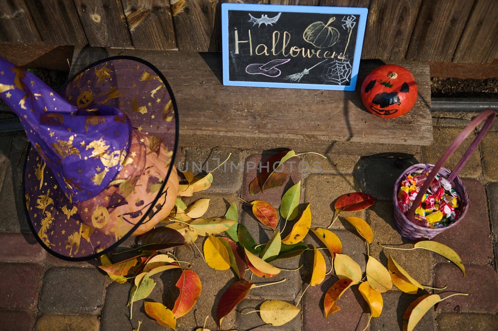 Top view: a blackboard with the inscription Halloween next to a pumpkin in wizard's hat on a wooden threshold with fallen yellow and red leaves in autumn. Porch or backyard decoration for Halloween