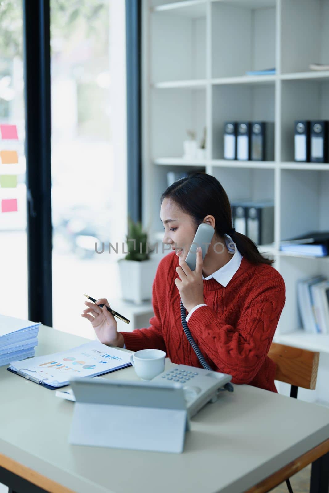 Portrait of a young Asian woman showing a smiling face as she uses her phone, computer and financial documents on her desk in the early morning hours by Manastrong