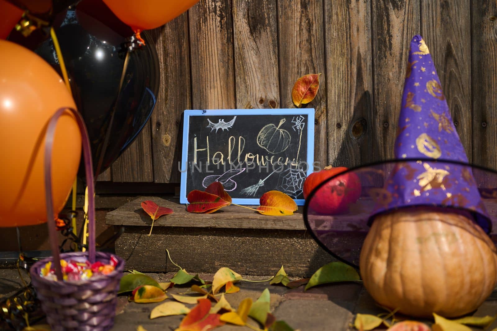 A wooden threshold or house porch, decorated for Halloween party with orange black air balloons, whole pumpkin in purple wizard's hat, fallen dry colorful autumn leaves and a violet basket with sweets