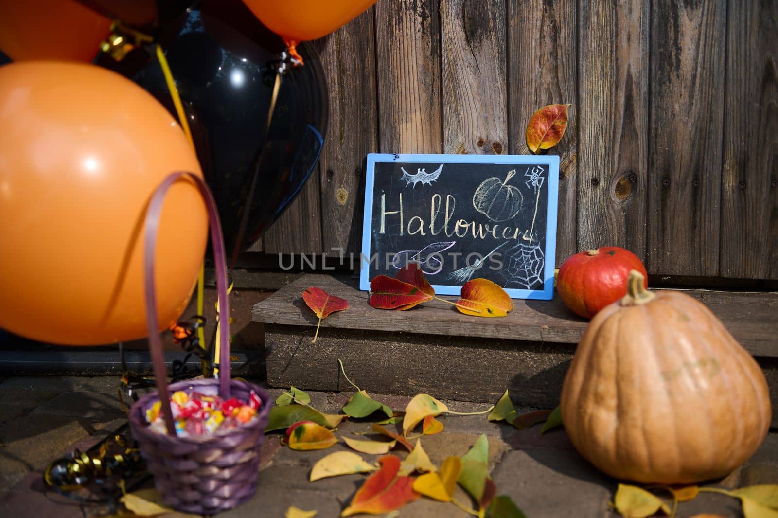 Pumpkin, candies, orange black balloons and blackboard on the wooden threshold with fallen autumn dry colorful leaves by artgf