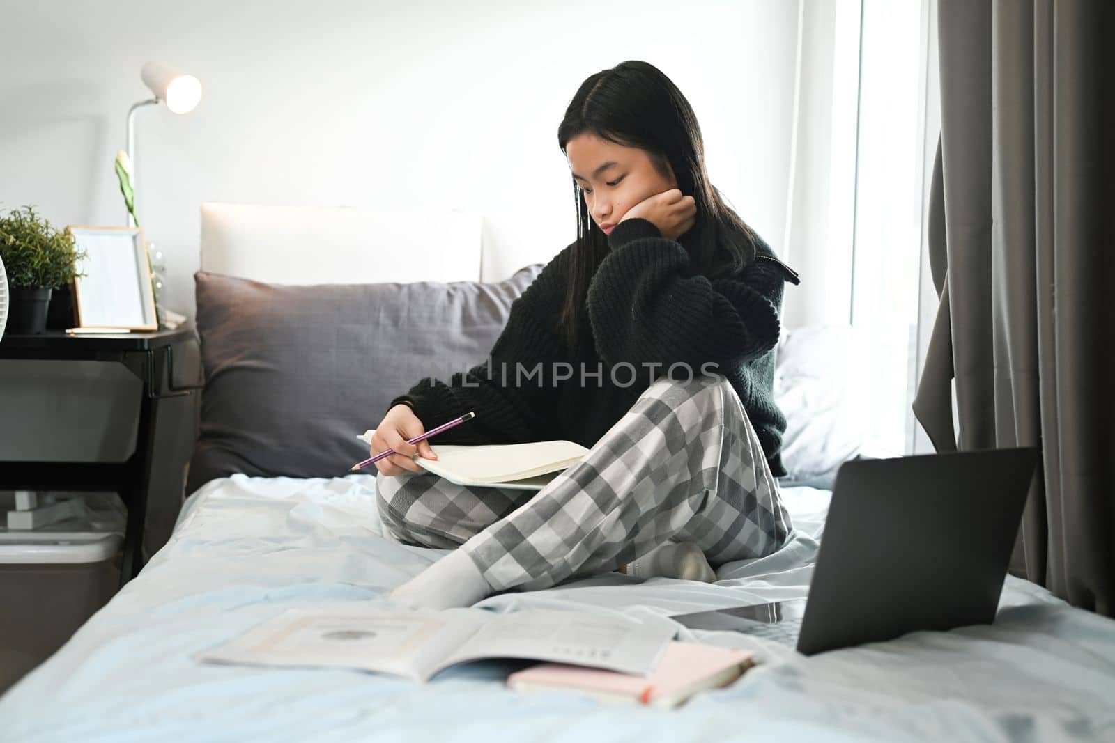 Teenage asian girl sitting on bed and doing homework. Distance education, studying online concept by prathanchorruangsak