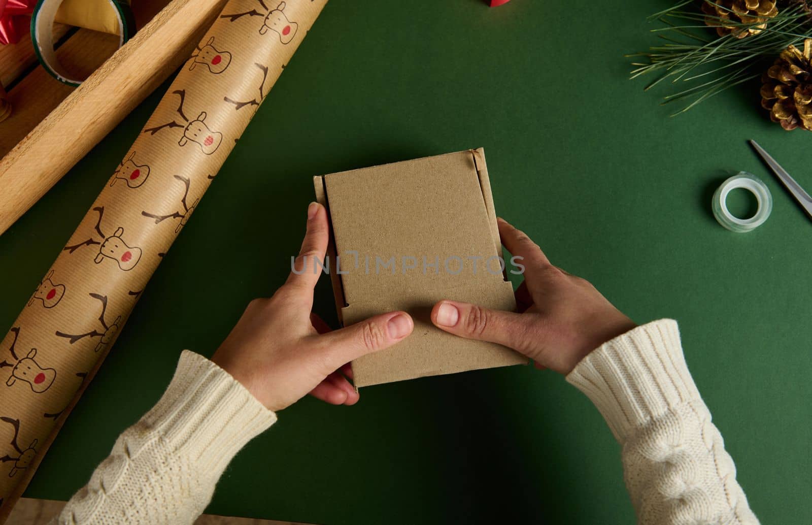 Top view female hands holding a cardboard box, packing gifts for Christmas or New Year event, on a green background with wrapping decorative meterials. Diy presents. Boxing Day. Handwork art craft