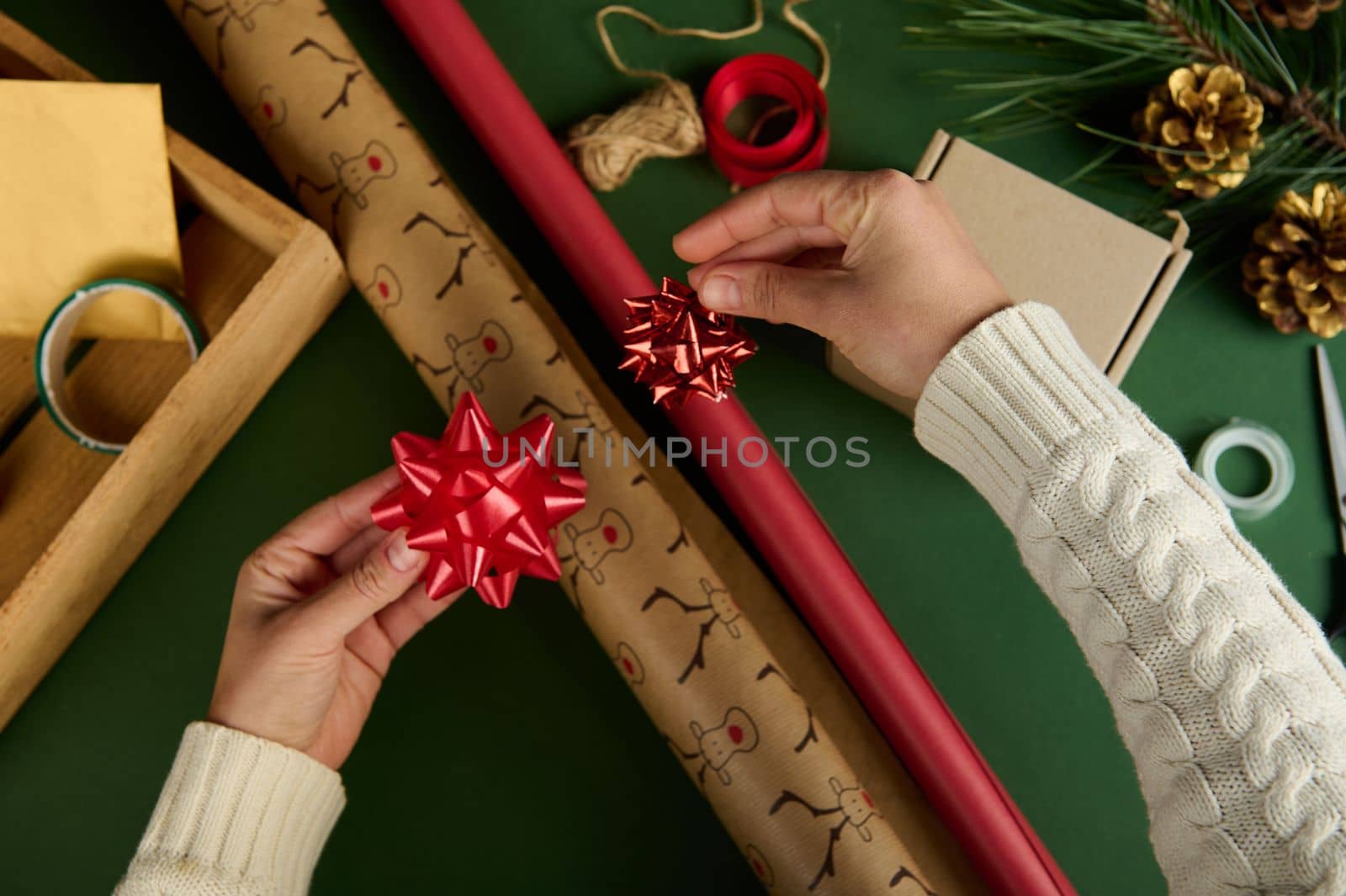 Top view woman's hands apply tied bows on wrapping paper with deer pattern and of red color, on a green surface with materials for packing Christmas and New Year gifts. Boxing Day. Handwork art. Diy