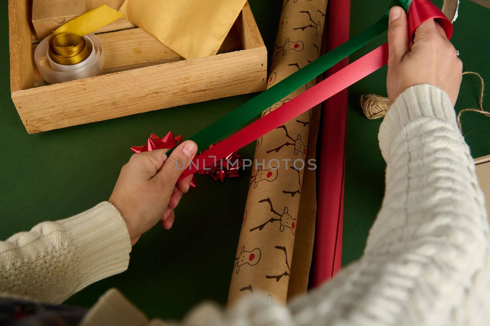 View from above of woman's hands holding red and green satin decorative tapes, over wrapping gift paper in art workshop by artgf