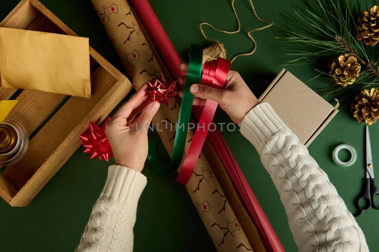 View from above of woman's hands holding red and green satin decorative tapes, a tied shiny bow over wrapping gift paper by artgf