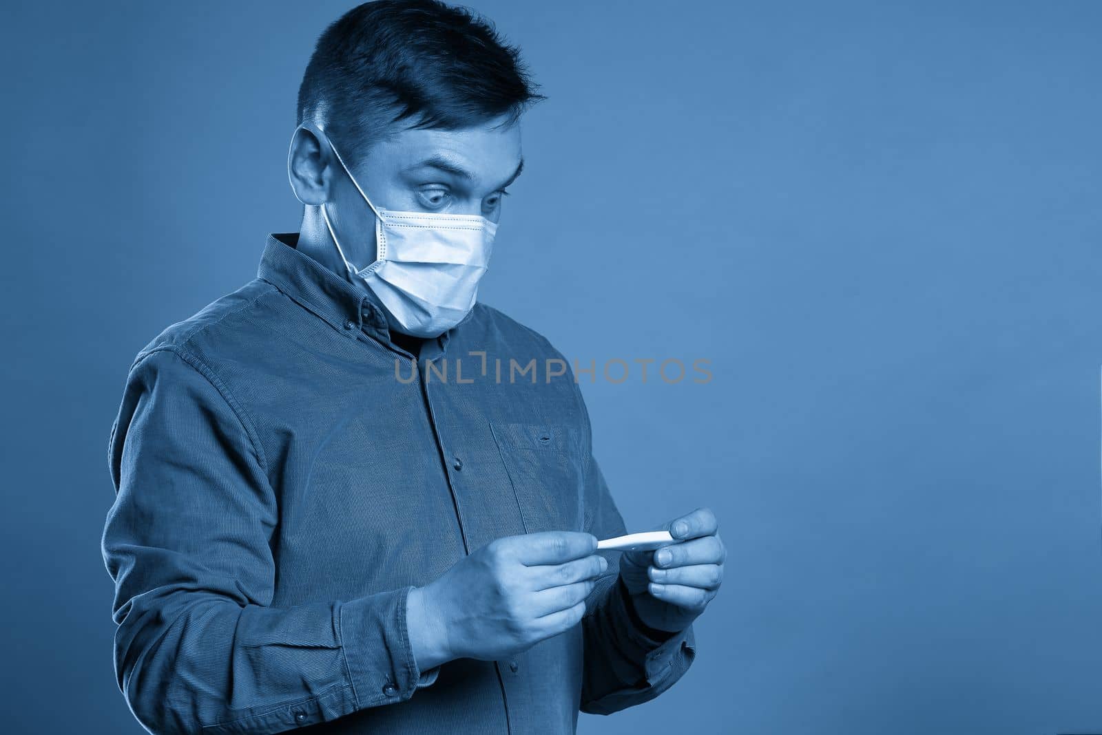 Young man in casual cloth wearing medical protective mask excited, stressed and frightened after read very high temperature result from digital thermometer