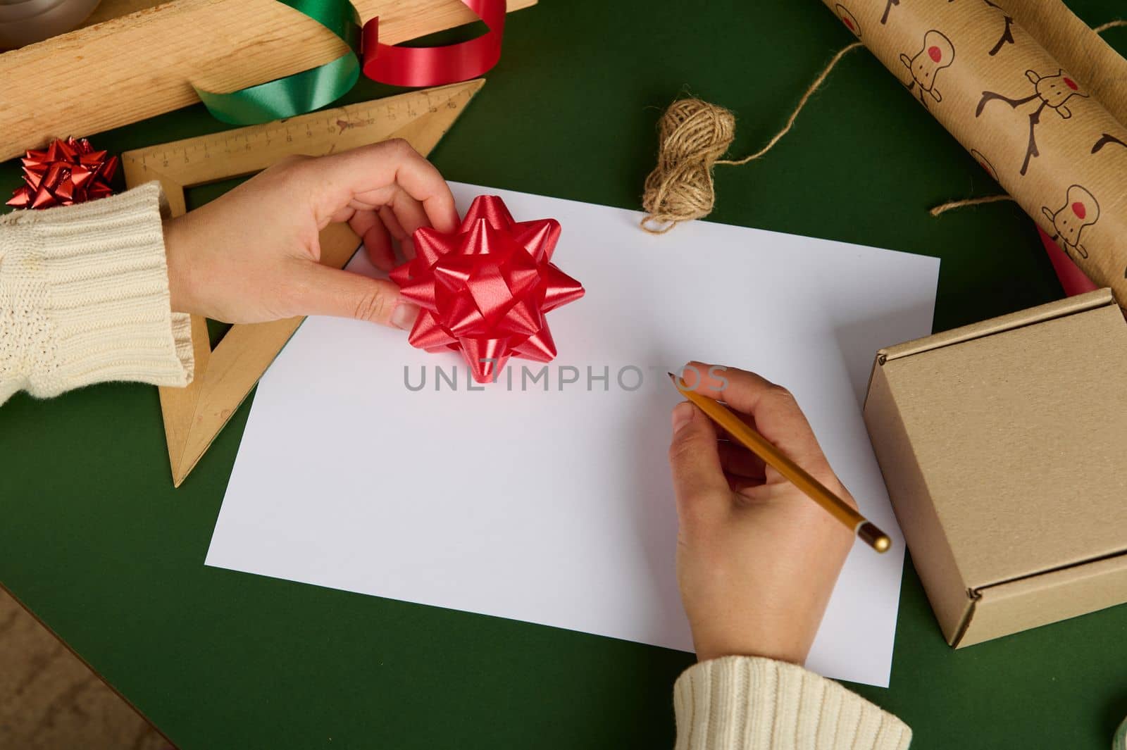 View from above. Female hand using pencil, draws, writes on a blank white paper sheet with copy space, holding a decorative red bow over a background with wrapping decorative materials. Packing gifts