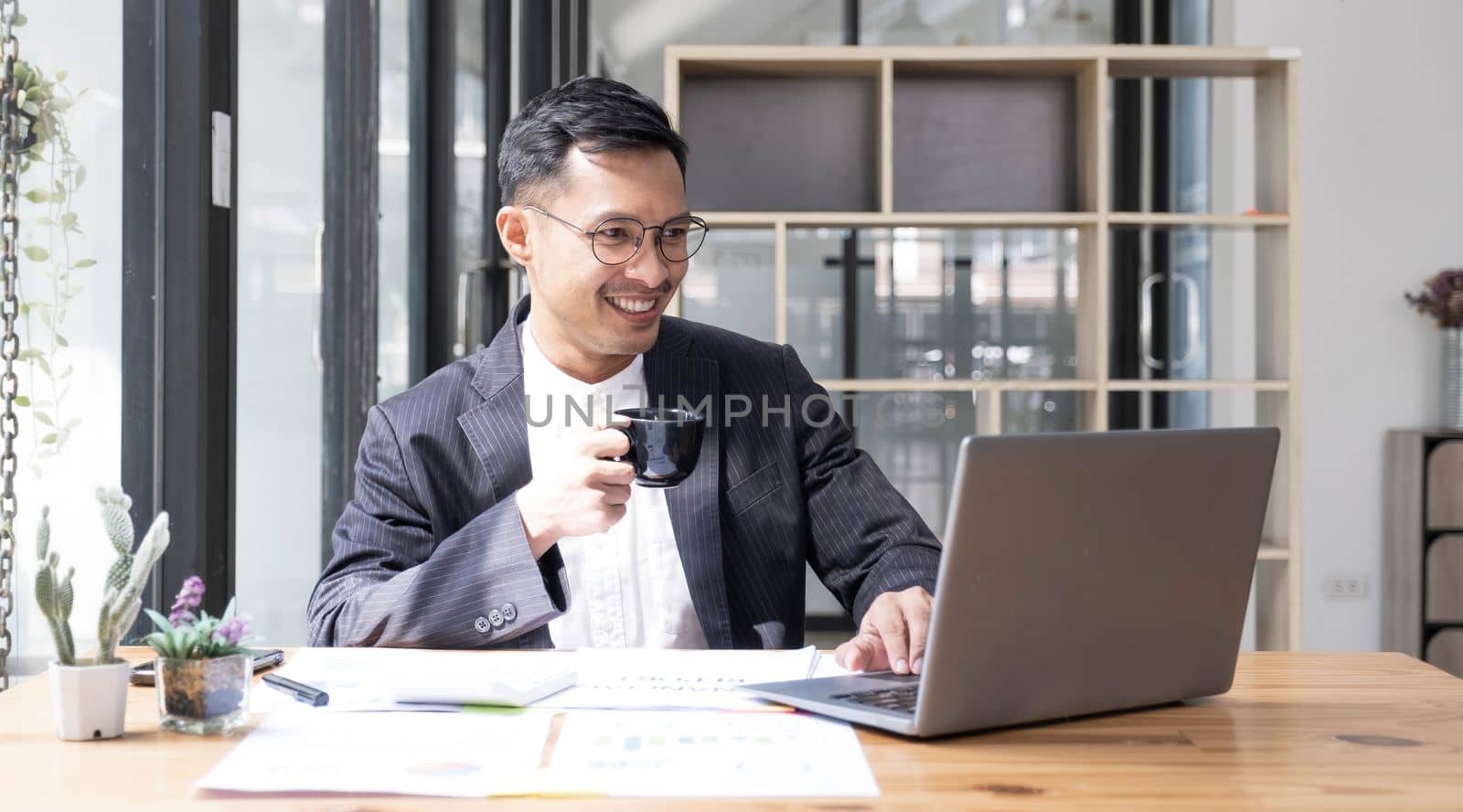 Young Asian businessman in suit and holding hot coffee cup and looking on laptop computer in office. Man in suit using laptop in well-lit workplace...
