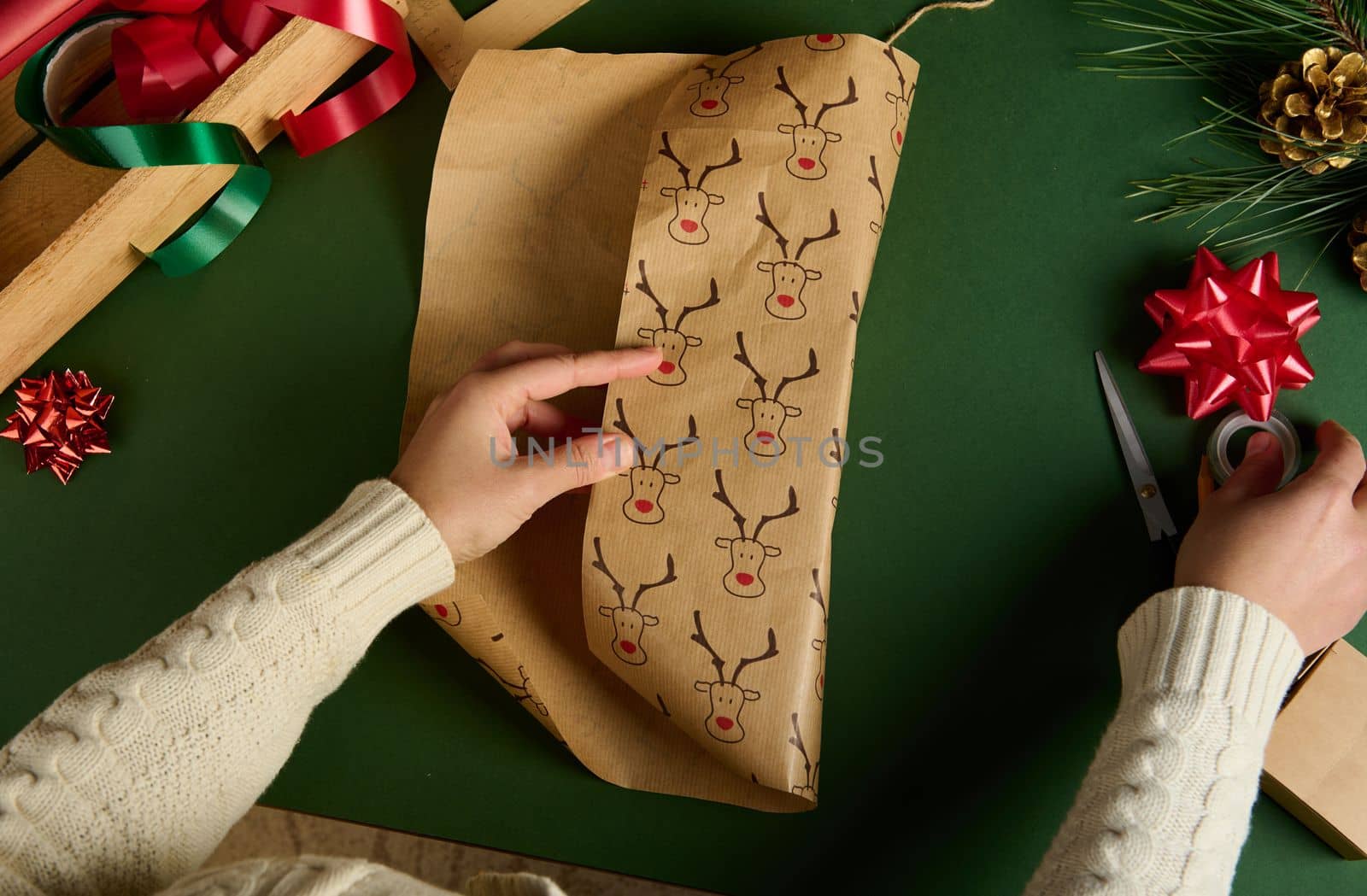 Top view of woman's hands wrapping a present for Christmas or New Year's events, using beautiful decorative paper with deer pattern, over green background. Boxing Day. Xmas preparations. Copy ad space