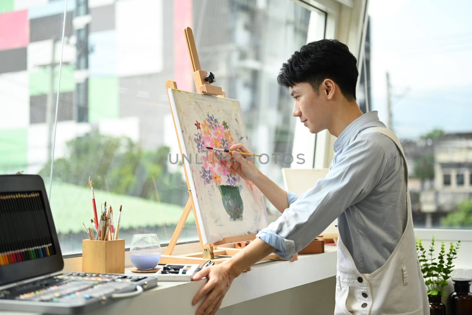Concentrated man painting picture on canvas near window at art workshop. Leisure activity, creative hobby and art concept. 