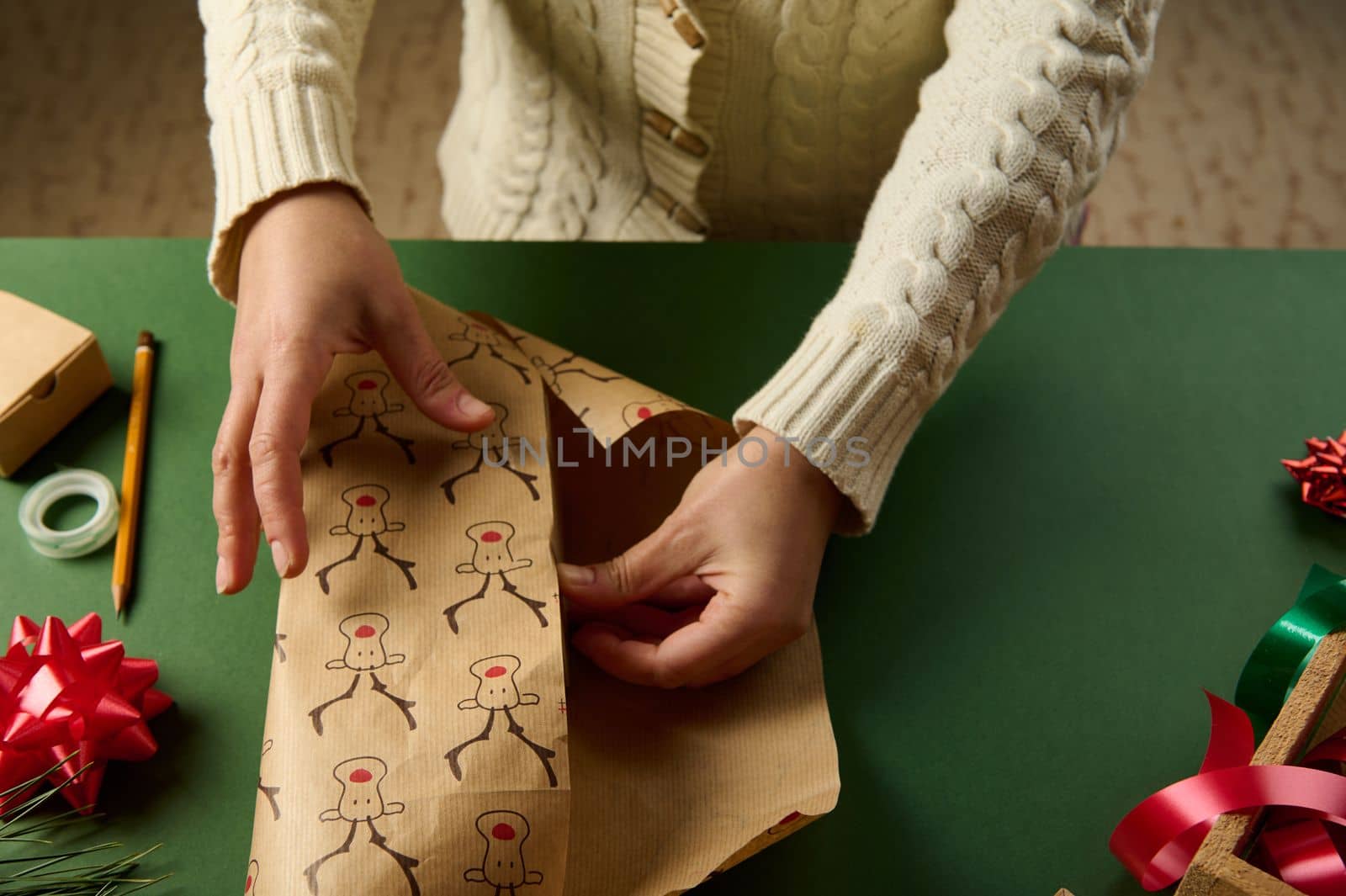 Close-up of a woman in beige sweater, wrapping a gift in a cute decorative paper with deer pattern, over a green background. Copy advertising space. Christmas and New Year's preparations. Boxing Day