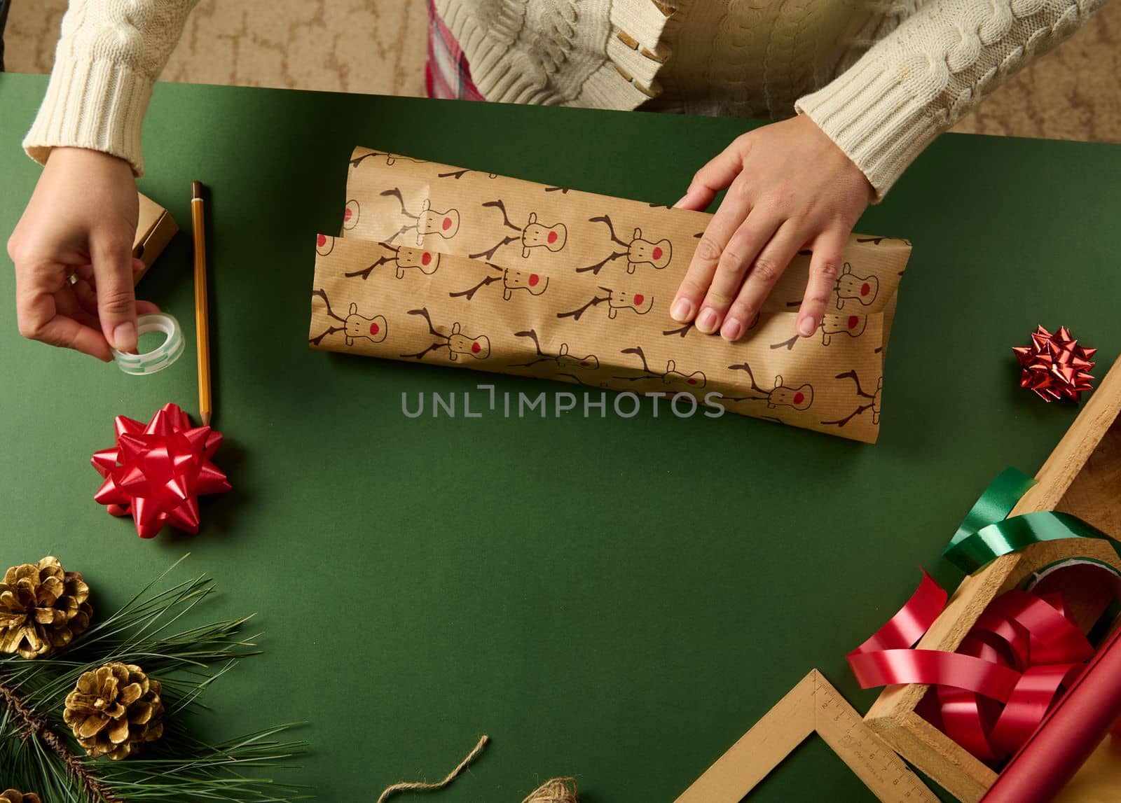 Close-up woman wrapping gift for Christmas in a paper with deer pattern, on a green background with golden pine cones as Xmas decoration, tied bow and decorative tapes in a wooden crate. Boxing Day