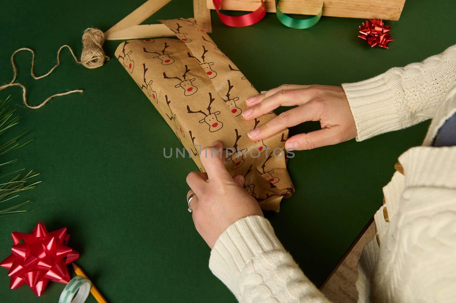 Top view of a woman in beige sweater, wrapping a gift in a cute decorative paper with deer pattern, over a green background. Copy advertising space. Christmas and New Year's preparations. Boxing Day
