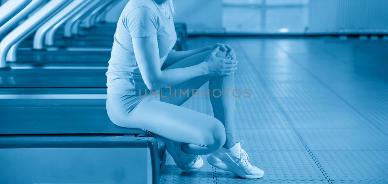 Young woman in sportswear having pain in her knee while training in gym, Girl sitting on a floor touching her knee in pain by Mariakray