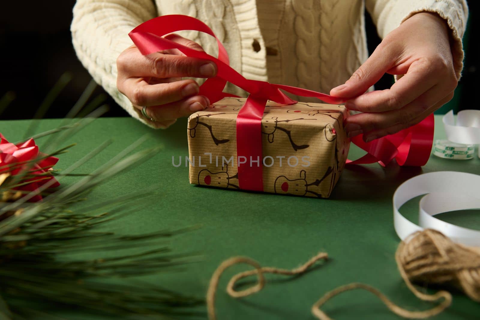 Close-up woman using a shiny red ribbon, ties up a gift wrapped in wrapping paper with deer pattern, over green background. Christmas and New Year's preparations. Boxing Day. New Year. Diy presents