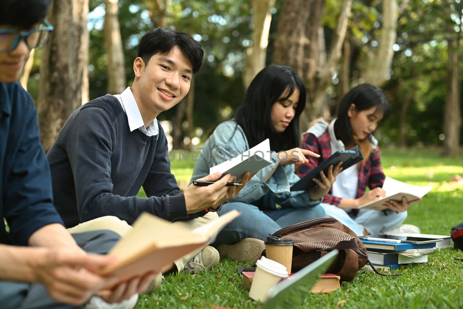 Asian student man reading books, preparing for exam on green lawn at university campus. Youth lifestyle and friendship concept.