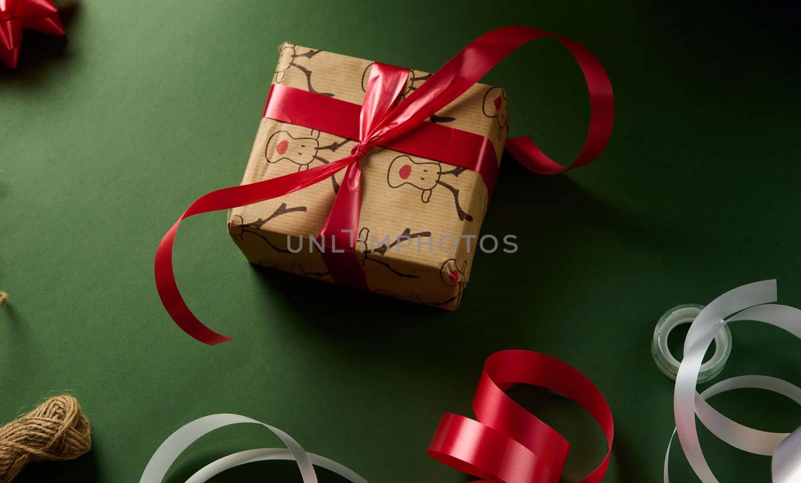 Top view of a cute Christmas gift box, wrapped in a decorative wrapping paper with deer pattern, tied up with a red shiny ribbon over green background. Boxing Day. Xmas and New Year preparations.