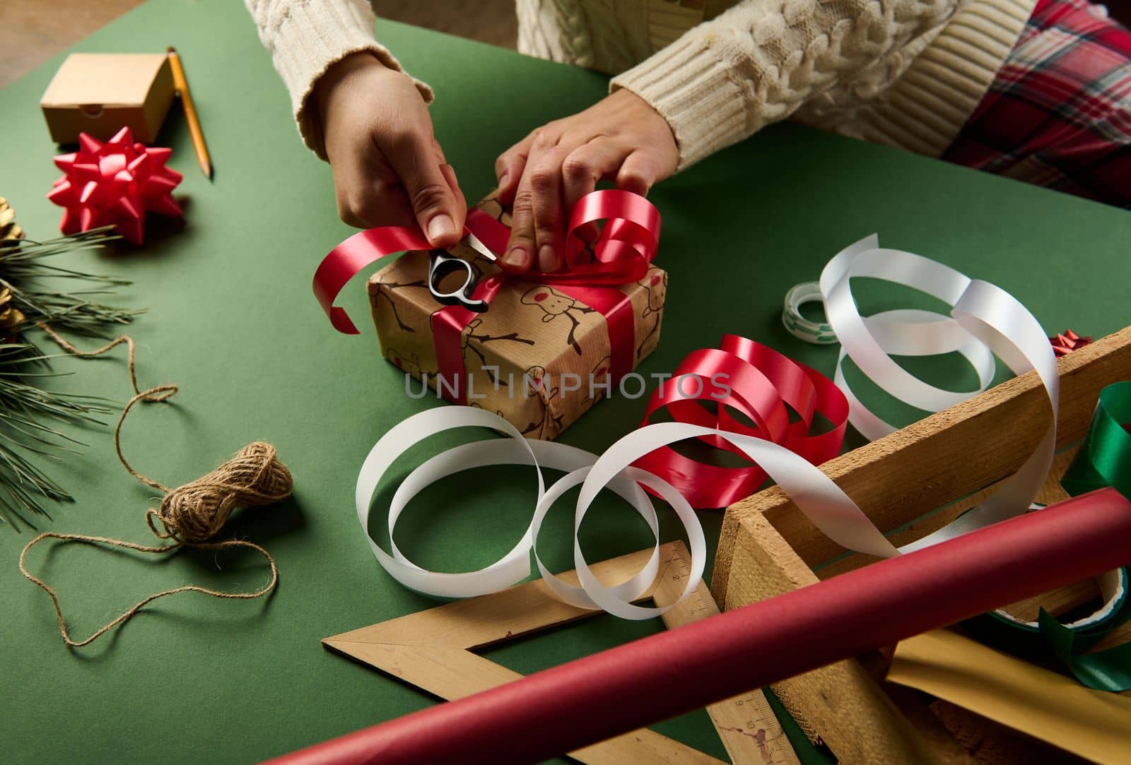 Close-up woman wraps Christmas gift box, tying up with a shiny red ribbon. Decorative tapes and bows, linen rope and ornaments on green background. New Year preparations. Packing presents. Boxing Day