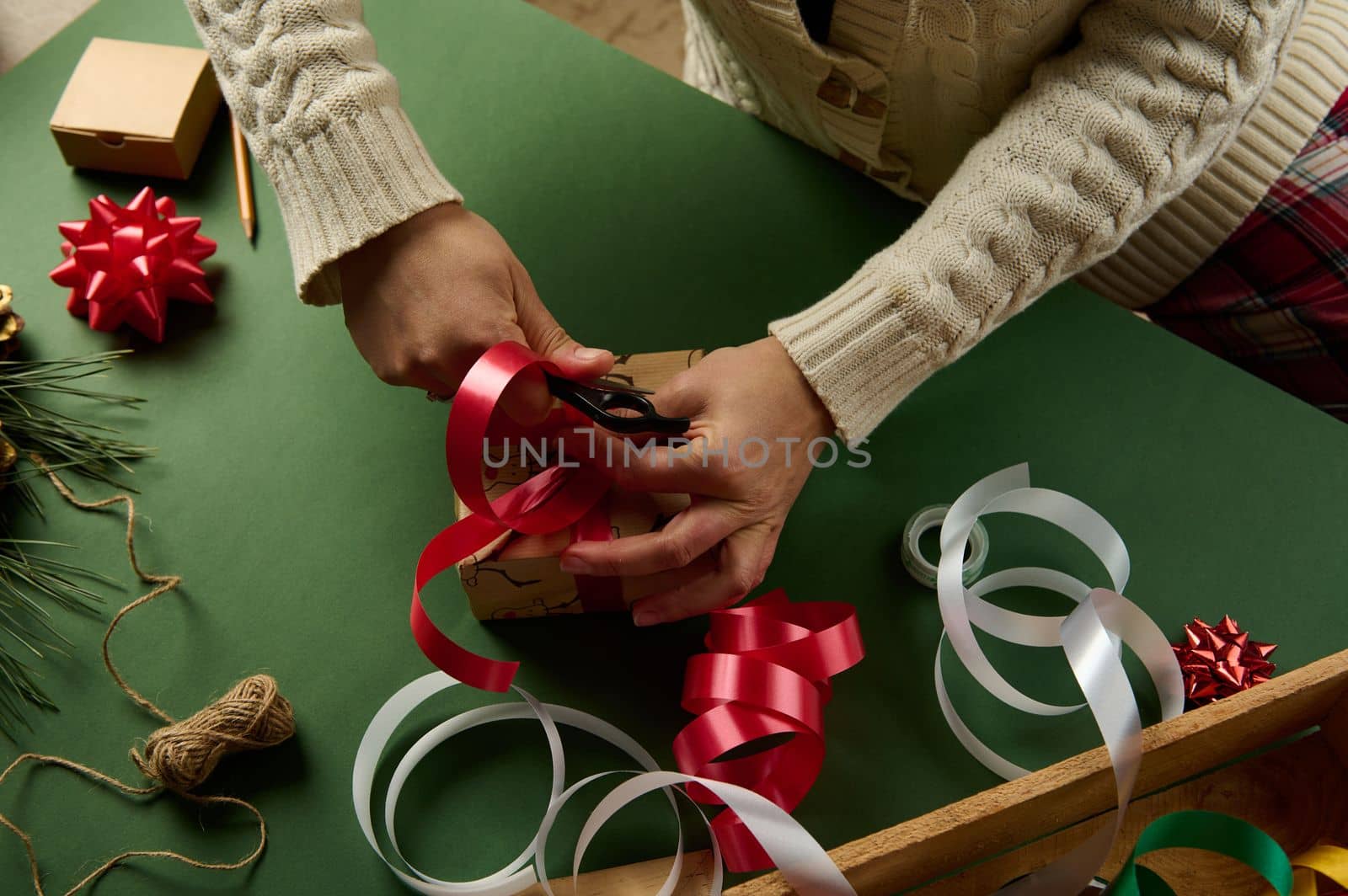 Top view woman in warm beige sweater, wrapping a Christmas or New Year's gift, tying a beautiful bow from a red shiny decorative ribbon. Boxing Day. Diy presents. Magical atmosphere of winter holidays