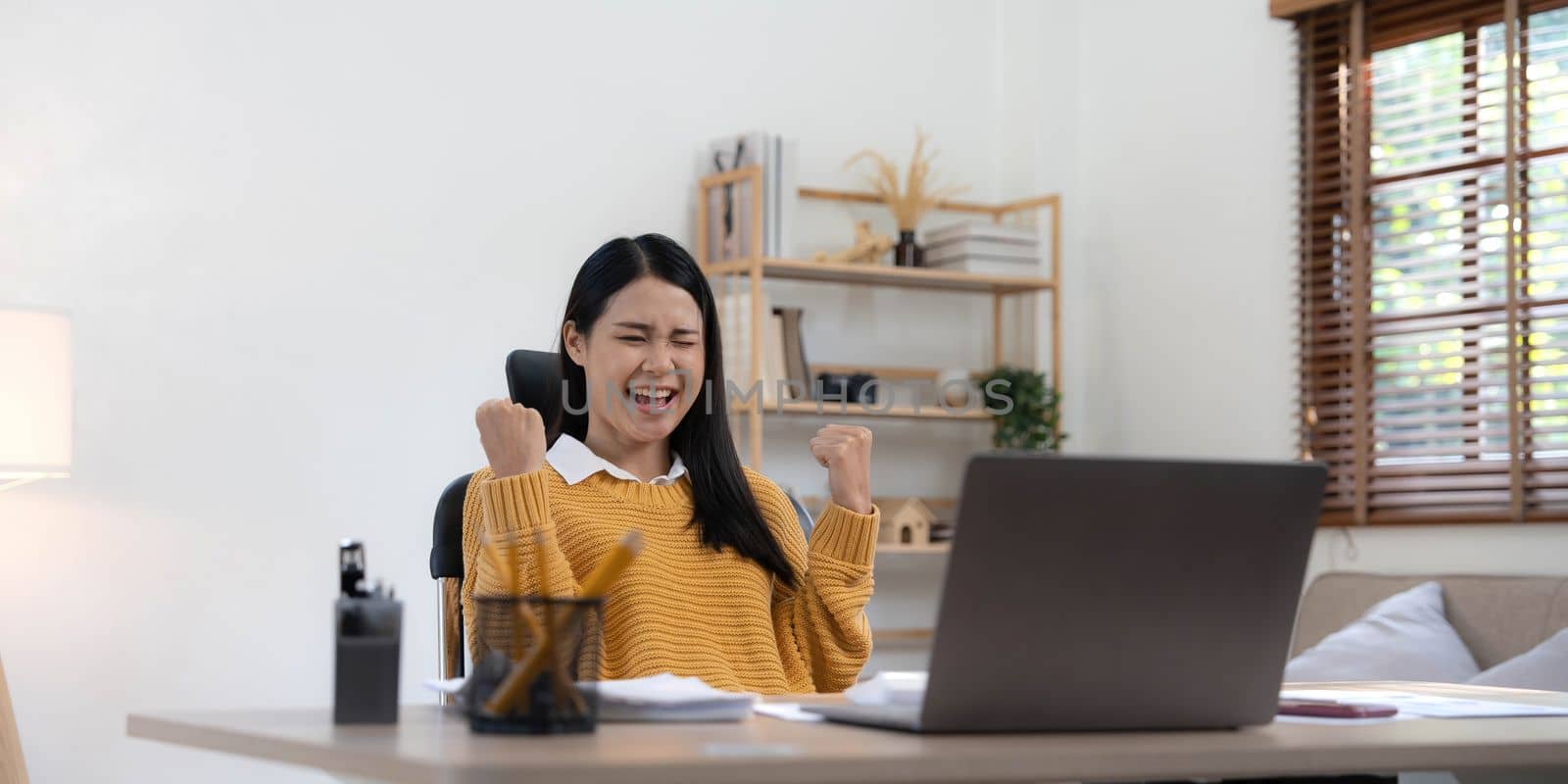 Excited happy woman looking at the laptop computer screen, celebrating an online win, overjoyed young asian female screaming with joy at home.