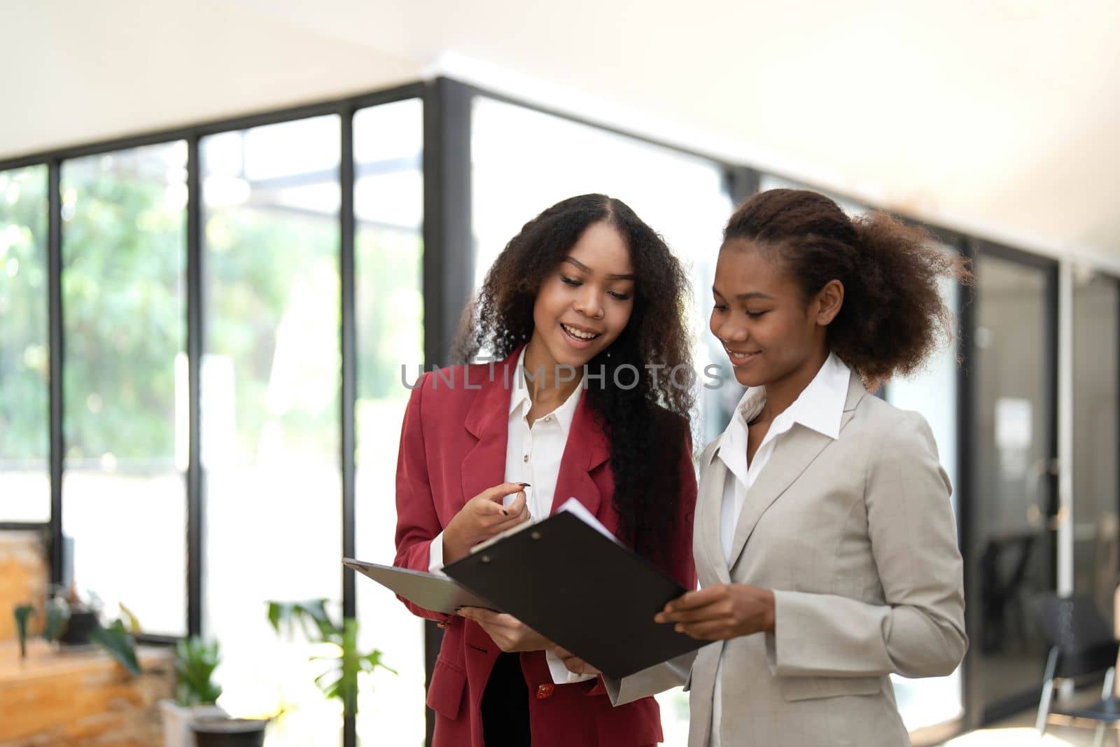 Smiling diverse colleagues businesswomen working on laptop together, looking at screen, stand at desk in office, employees discussing project strategy, sharing ideas by wichayada