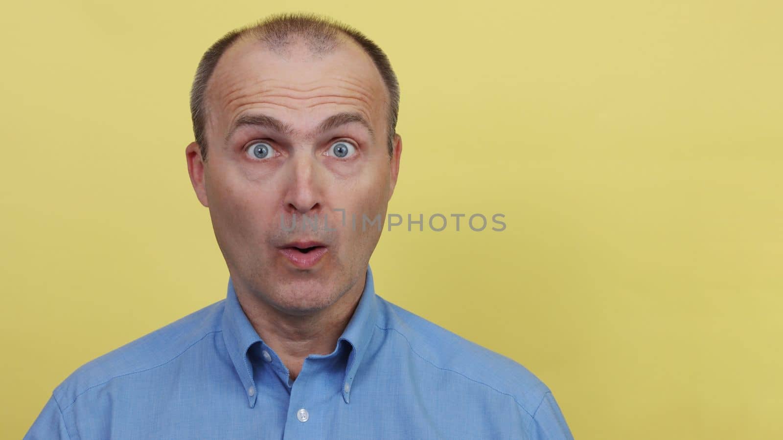 Portrait of a surprised handsome man 45-55 years old in a blue shirt looks at the camera.A man in a blue shirt on a yellow background.