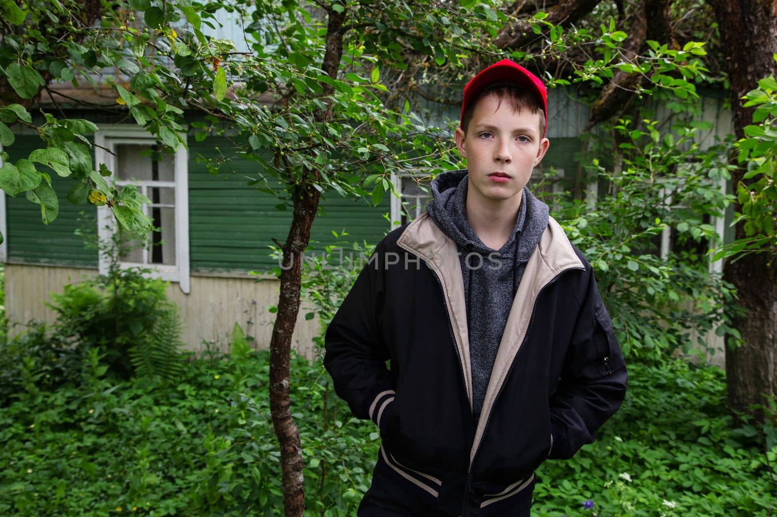 A boy in a demi-season jacket looks strictly at the camera against the backdrop of a village house. Rest of the boy in the village. boy on the background of foliage and a country house