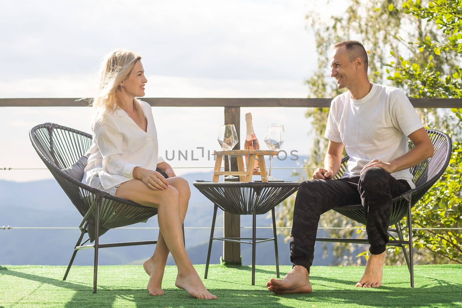 Happy couple sit on wicker chairs at outdoor terrace in the mountains and drink wine. Romantic time together