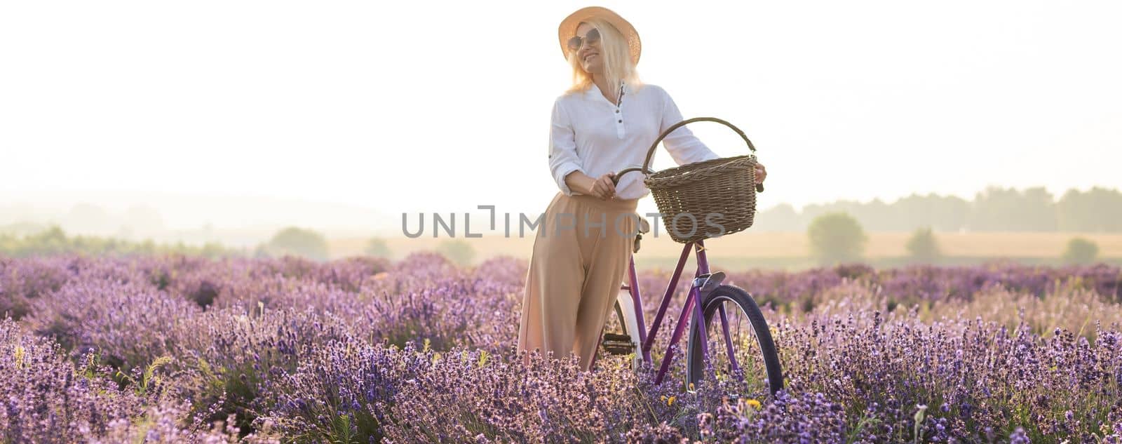 Beautiful young healthy woman with a white dress running joyfully through a lavender field holding a straw hat under the rays of the setting sun.