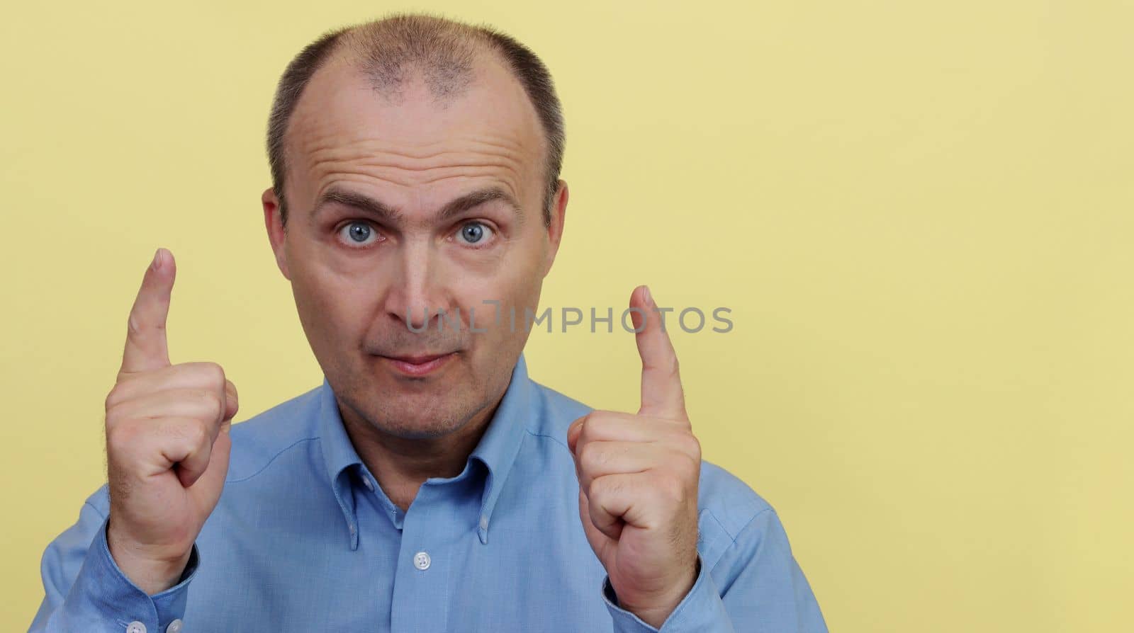 A man 45-55 years old in a blue shirt on a yellow background raised two hands with an index finger.