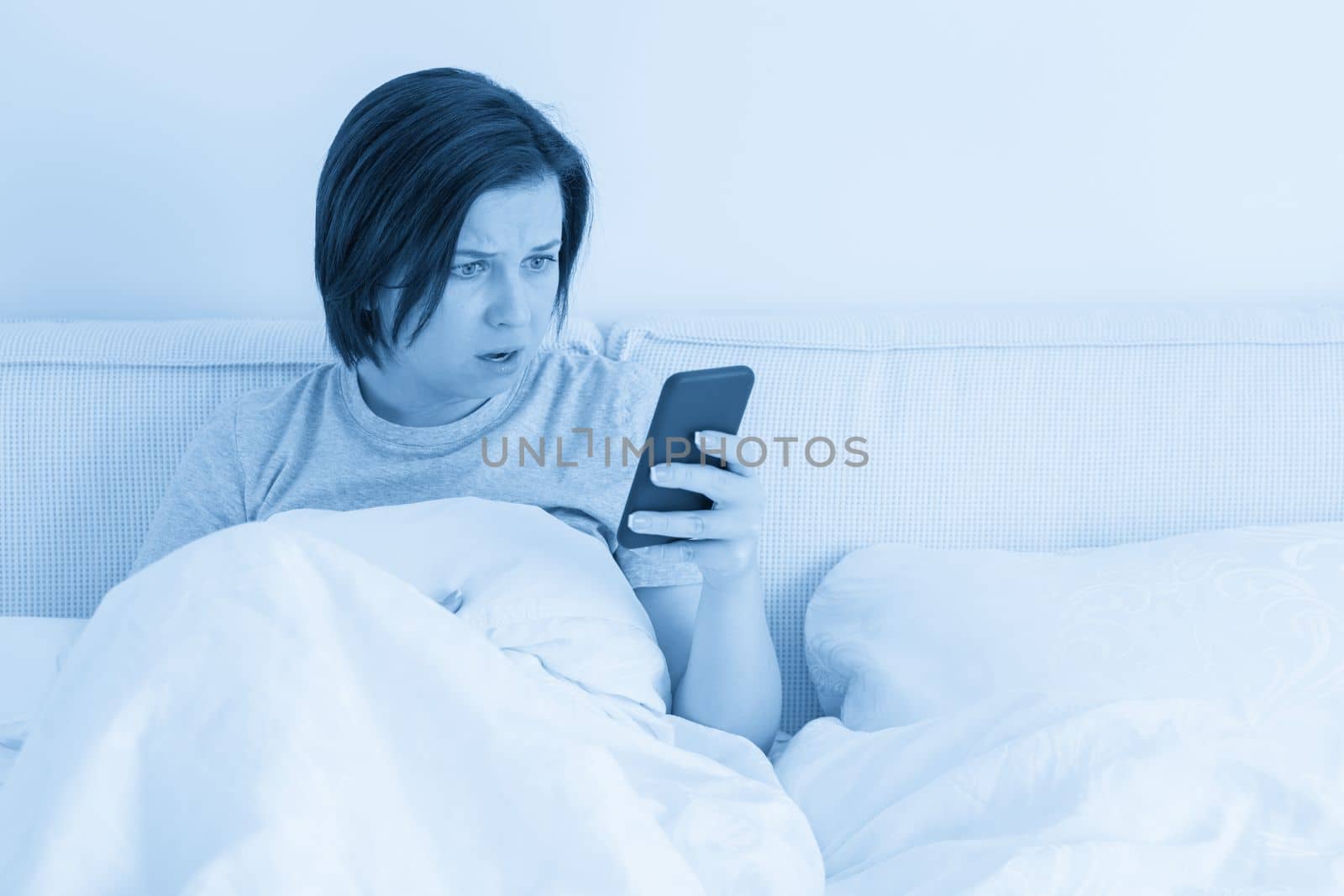 Woman using smartphone as she sits in bed covered with a duvet