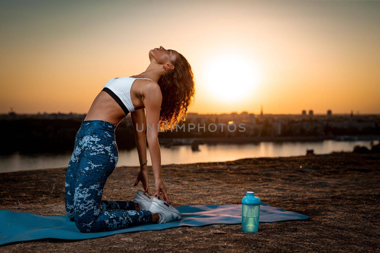 Young urban woman doing stretching exercises by the river in a sunset.