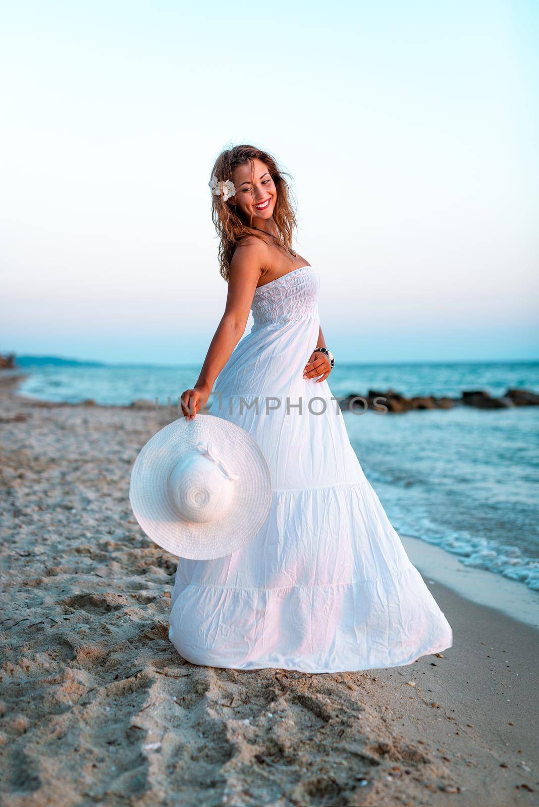 Beautiful young smiling woman in white dress walking on the beach and holding white summer hat.