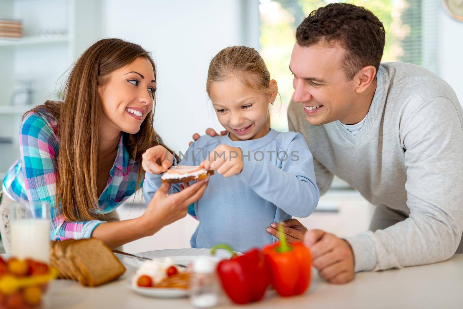 Beautiful young family preparing healthy meal for the breakfast in the domestic kitchen. The mother learns the little girl how to make the sandwich and father is looking what she is doing.