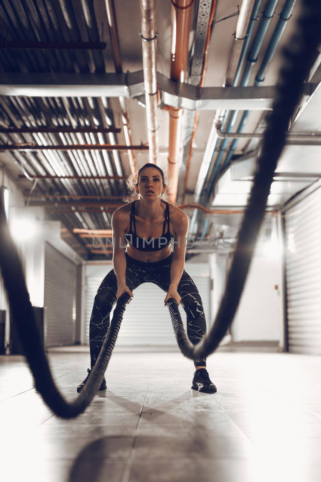 Muscular young woman exercising with ropes at the garage gym. Selective focus.