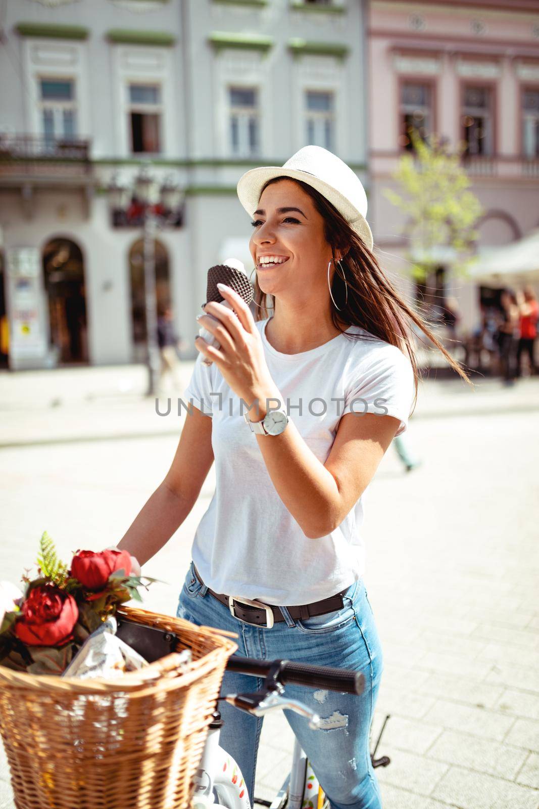 Smiling young woman with ice cream, taking break on a city square on a sunny day, beside the bike with flower basket.
